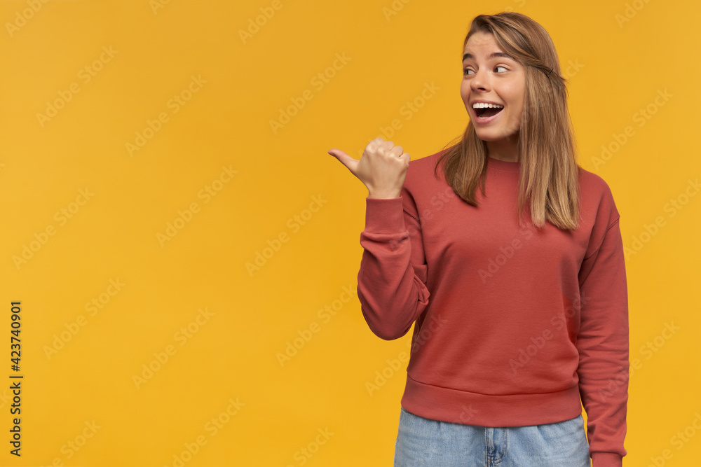 lovely young beautiful woman smiles broadly and indicates with finger to copyspace posing over orange background with happy facial expression wears red pullover and blue denim pants