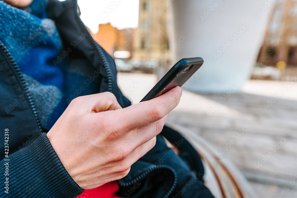 A young man in a jacket sits on a bench near a business building and uses a phone