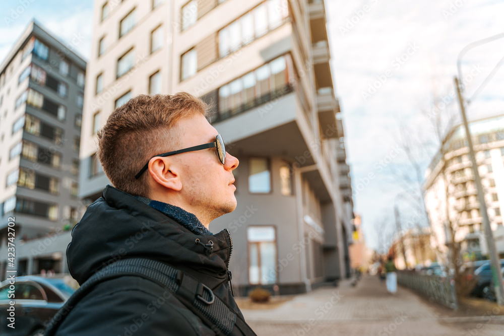 A young man in a jacket and sunglasses stands in the financial district near the business buildings A young man in a jacket and sunglasses stands in the financial district near the business buildings