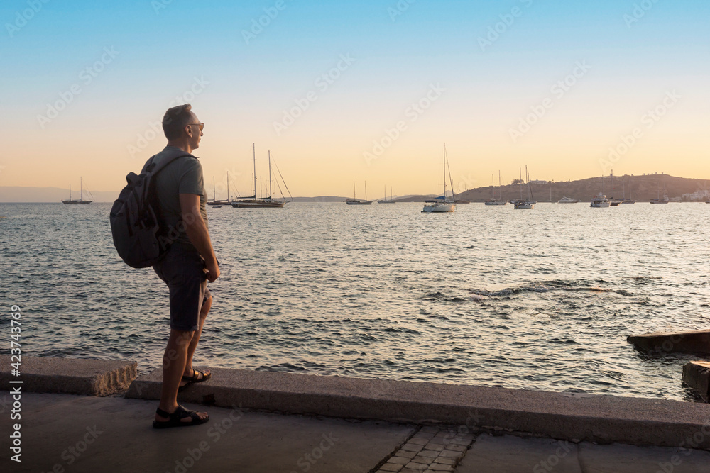 Tourist with backpack standing on a seafront of Bodrum and enjoing beautiful view at sunset. Selective focus.