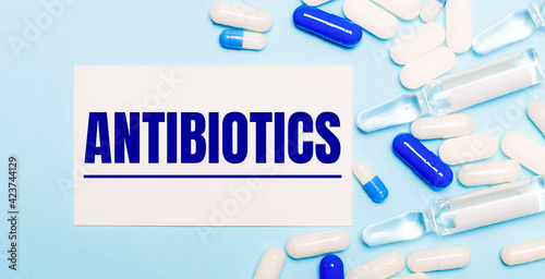 Pills, ampoules and a white card with the text ANTIBIOTICS on a light blue background. Medical concept