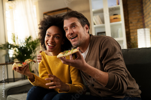 Cheerful young couple sitting on sofa at home. Happy woman and man eating pizza while watching a movie