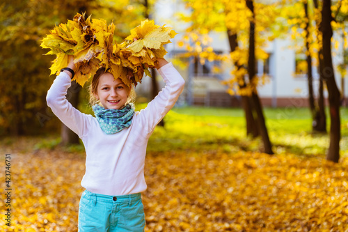 Cute blond caucasian girl in head wreath from yellow maple leaves in autumn park. A little girl in an autumn sweater playing with maple wreath at sunny warm day, outdoor.