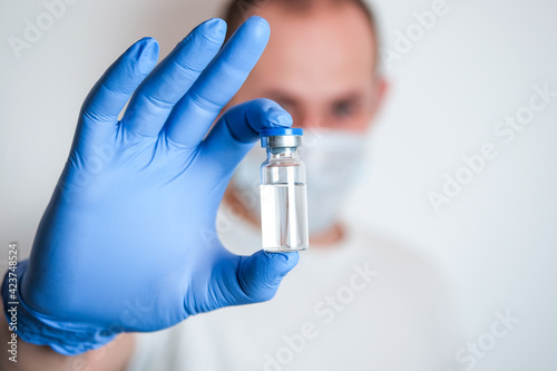 A hand in blue protective glove holding an ampoule with a liquid medicine with a blurred man on the background. Vaccination concept. Injection for prevention and health care.