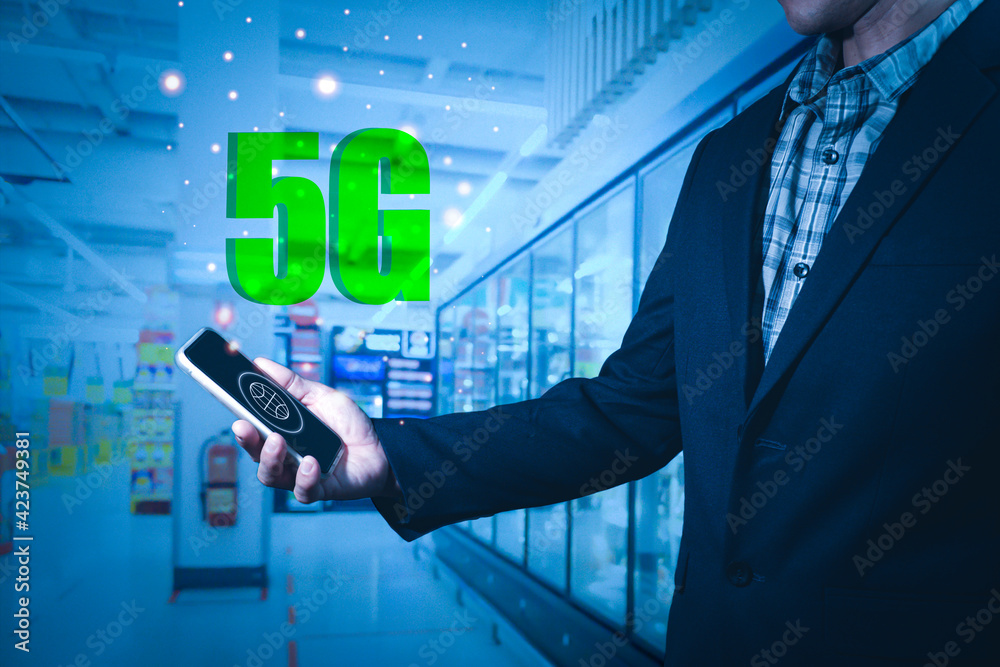 A man wearing a suit holds a mobile phone on the screen in a world shape with 5G letters floating on the screen. The background is layered and the shelves are blurred. A man wearing a suit holds a mobile phone on the screen in a world shape with 5G letters floating on the screen. The background is layered and the shelves are blurred.