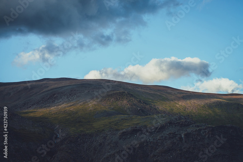 Minimalist alpine landscape with sunlight on mountain silhouette under blue sky with clouds. Scenic view to large mountain under cloudy sky. Minimal nature background with light and dark on mountain.