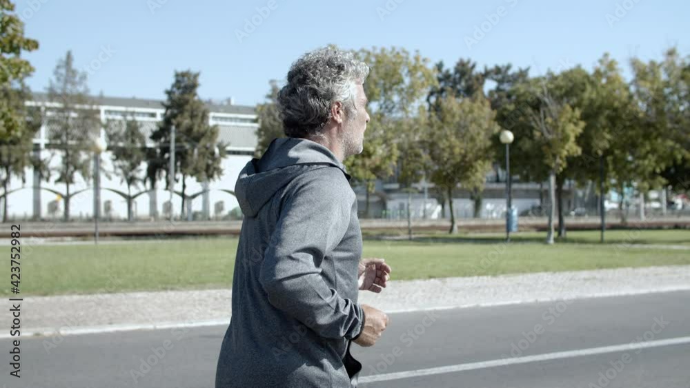 Older male runner jogging outside. Active elderly man in sporty clothes keeping fit, running on promenade. Dolly shot. Elderly age and lifestyle concept Older male runner jogging outside. Active elderly man in sporty clothes keeping fit, running on promenade. Dolly shot. Elderly age and lifestyle concept