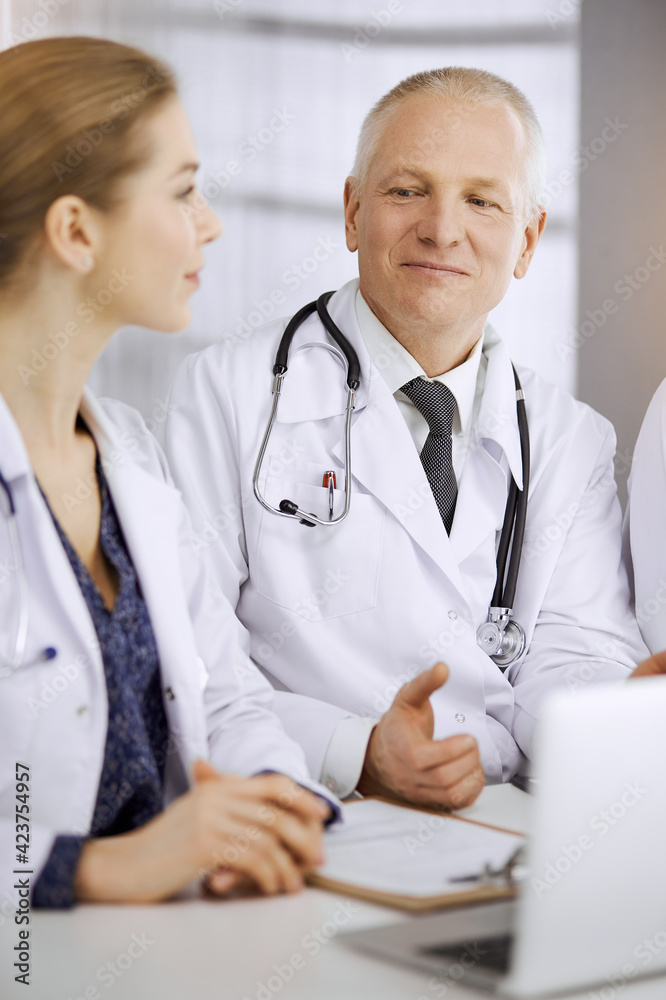 Elderly doctor and his female colleague discussing current disease therapy while sitting at working place in sunny clinic. Teamwork in medicine Elderly doctor and his female colleague discussing current disease therapy while sitting at working place in sunny clinic. Teamwork in medicine