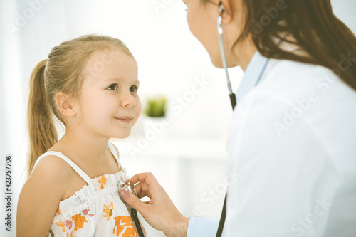 Doctor examining a child by stethoscope. Happy smiling girl patient dressed in bright color dress is at usual medical inspection