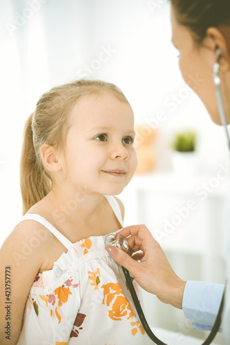 Doctor examining a child by stethoscope. Happy smiling girl patient dressed in bright color dress is at usual medical inspection