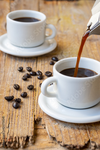 Top view of coffee pot serving in white cup, on rustic wooden table with full cup and coffee beans, selective focus, vertical