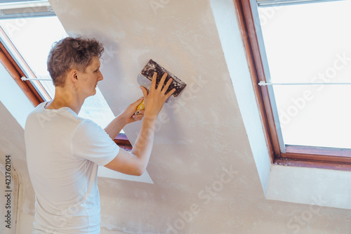 A young man scratching with a spatula and removing old wallpaper on a wall of a room. Do it yourself concept. Home renovation. Selective focus, copy space. photo