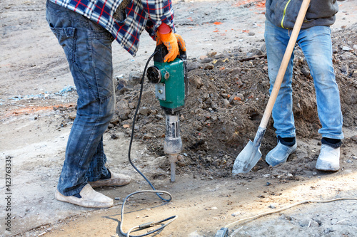 Builders at a construction site are loosening the dense soil with an electric jackhammer and shovel.