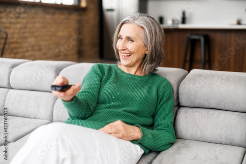 Cheerful senior woman enjoys evening with a TV shows, series. Smiling mature woman sits on the sofa with TV remote controller and switching channels