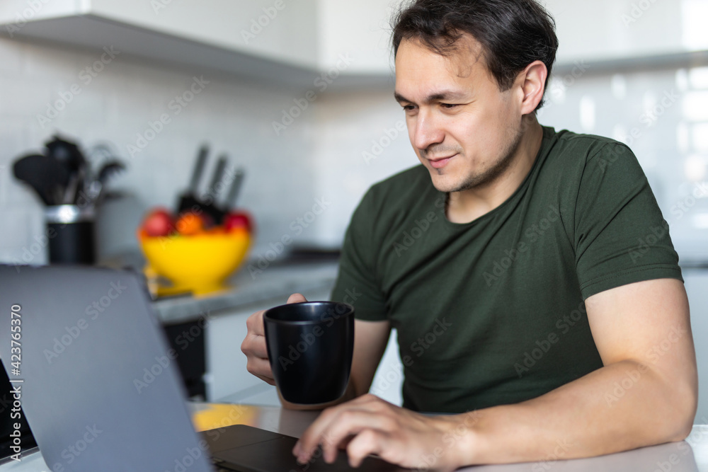 Young man working on his laptop while drinking coffee in the kitchen Young man working on his laptop while drinking coffee in the kitchen