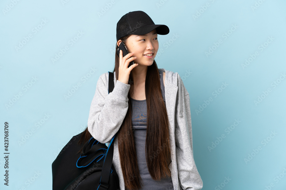 Young sport Chinese woman with sport bag over isolated blue background keeping a conversation with the mobile phone with someone Young sport Chinese woman with sport bag over isolated blue background keeping a conversation with the mobile phone with someone