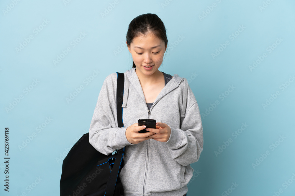 Young sport Chinese woman with sport bag over isolated blue background sending a message with the mobile Young sport Chinese woman with sport bag over isolated blue background sending a message with the mobile