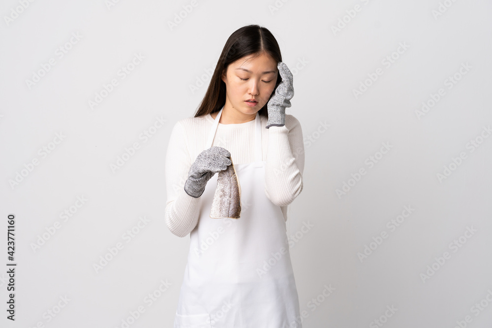 Chinese fishmonger wearing an apron and holding a raw fish over isolated white background with headache Chinese fishmonger wearing an apron and holding a raw fish over isolated white background with headache