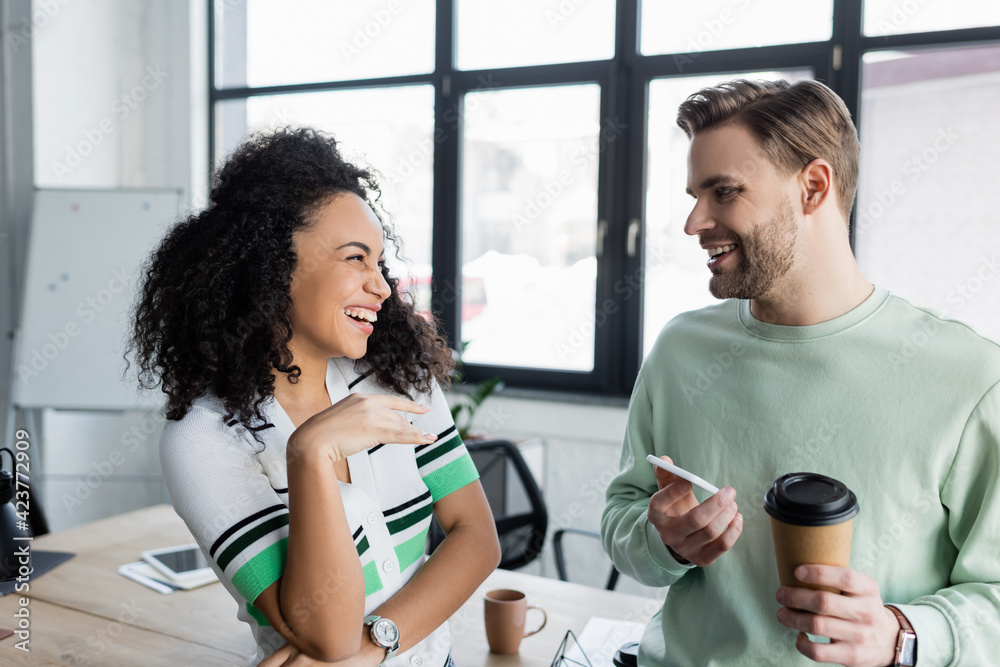 Fototapeta premium happy businessman holding coffee and smartphone while talking to laughing african american colleague