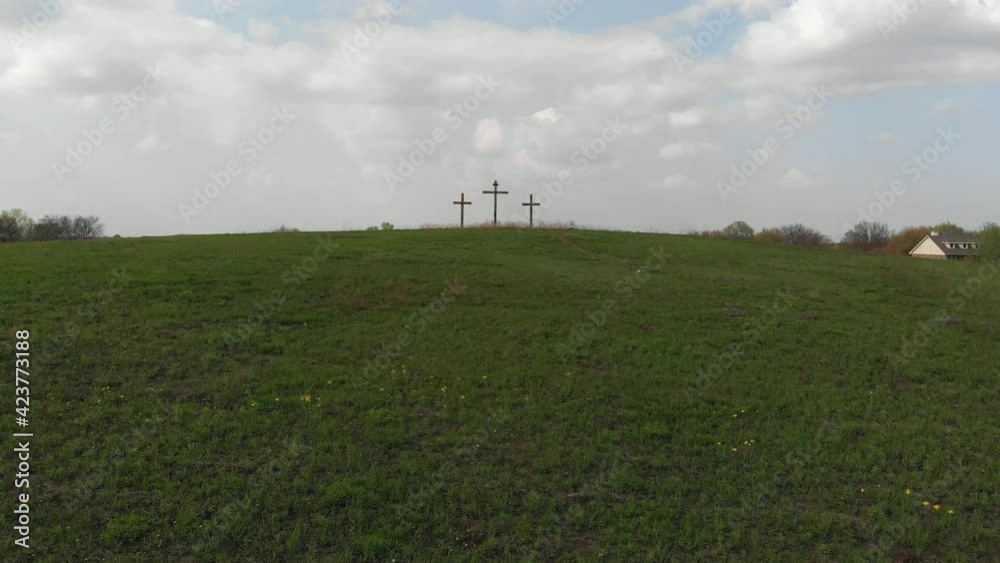 Three Easter crosses on a yellow flower dappled grassy hill overlooking a small town and a public road with cars traveling past as recorded by drone Three Easter crosses on a yellow flower dappled grassy hill overlooking a small town and a public road with cars traveling past as recorded by drone