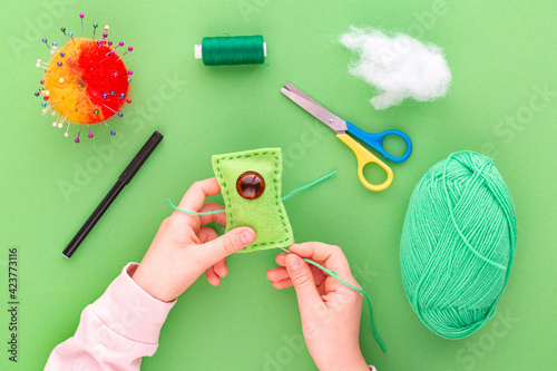 Instruction step by step, step nine. The hands of a child in the process of sewing a toy from felt, a green one-eyed monster. Green background, flat lay. Hobbies, handicrafts, fine motor skills.