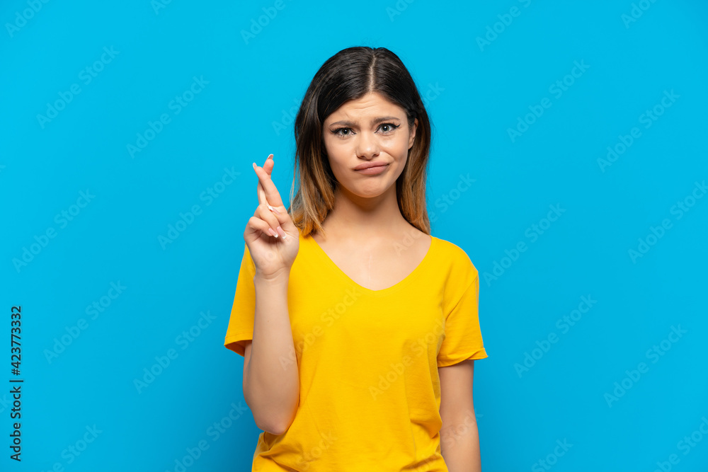 Young Russian girl isolated on blue background with fingers crossing and wishing the best Young Russian girl isolated on blue background with fingers crossing and wishing the best