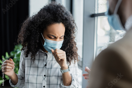 diseased african american businesswoman in medical mask coughing near coworker on blurred foreground