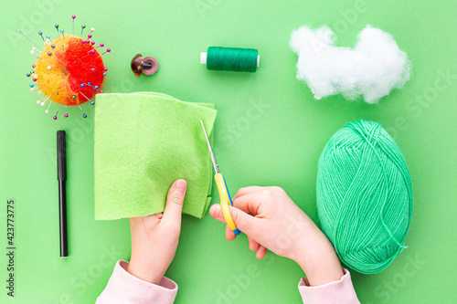 Instruction step by step, step one. The hands of a child in the process of sewing a toy from felt, a green one-eyed monster. Green background, flat lay. Hobbies, handicrafts, fine motor skills.