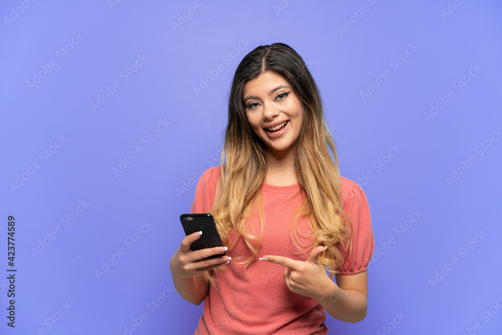 Young Russian girl isolated on blue background using mobile phone and pointing it Young Russian girl isolated on blue background using mobile phone and pointing it