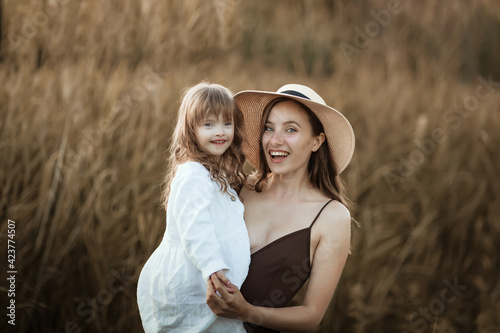 Charming Caucasian young mom holds her daughter with Down syndrome, beautiful special motherhood, mother with daughter hugging in summer in park, chocolate brown toning, evening light