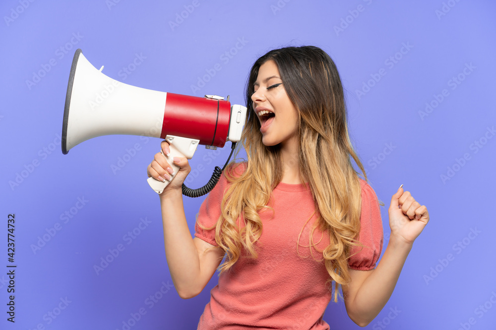 Young Russian girl isolated on blue background shouting through a megaphone to announce something in lateral position Young Russian girl isolated on blue background shouting through a megaphone to announce something in lateral position