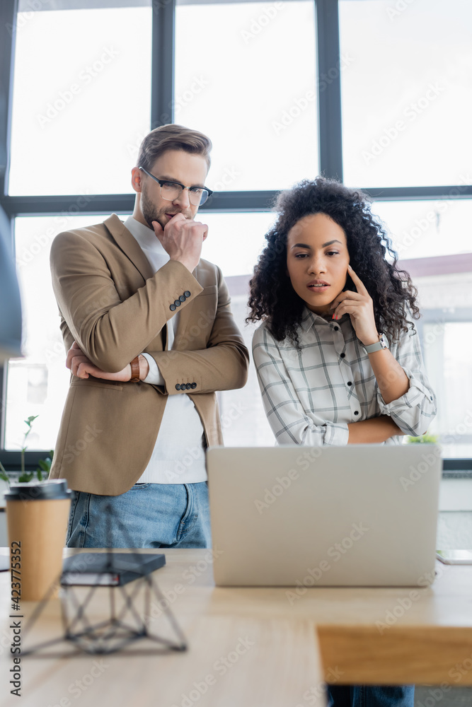 Focused african american businesswoman looking at laptop near colleague in office Focused african american businesswoman looking at laptop near colleague in office