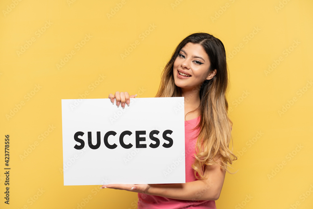 Young Russian girl isolated on yellow background holding a placard with text SUCCESS with happy expression Young Russian girl isolated on yellow background holding a placard with text SUCCESS with happy expression