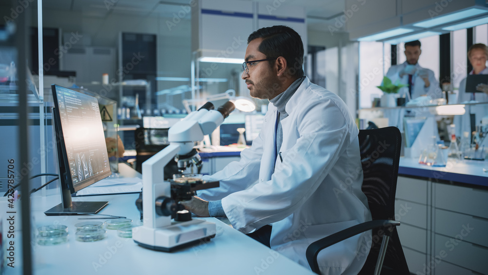 Medical Research Scientist Conducts DNA Experiments With Use of Microscope, Writes Results on Desktop Computer in Biological Science Laboratory. Diverse Multiethnic Lab with Engineers in White Coats. Medical Research Scientist Conducts DNA Experiments With Use of Microscope, Writes Results on Desktop Computer in Biological Science Laboratory. Diverse Multiethnic Lab with Engineers in White Coats.