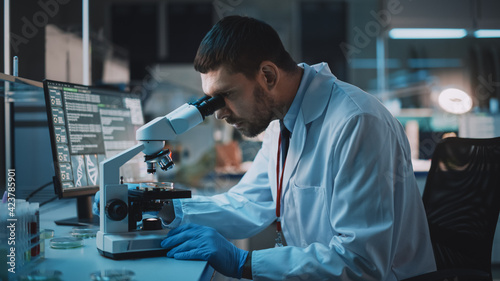 Medical Research Scientist Conducts DNA Experiments Under Digital Microscope in a Biological Applied Science Laboratory. Handsome Caucasian Lab Engineer in White Coat Working on Vaccine and Medicine.