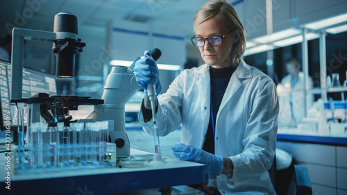 Female Research Scientist is Wearing Glasses and Using Micropipette to Extract a Sample on a Microscope Slide to Make Analysis of it. Scientists Workingin Modern Laboratory with Help of Technologies.