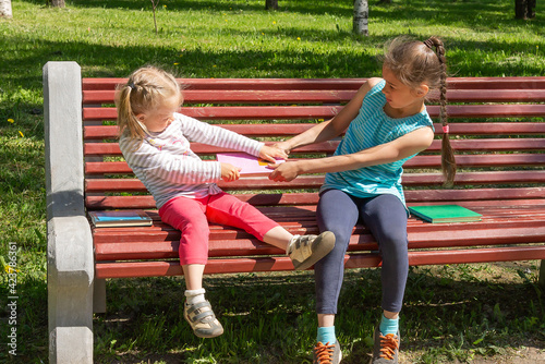 Two kids quarrel on a bench in the park. Two little girls do not want to share and are fighting over a book