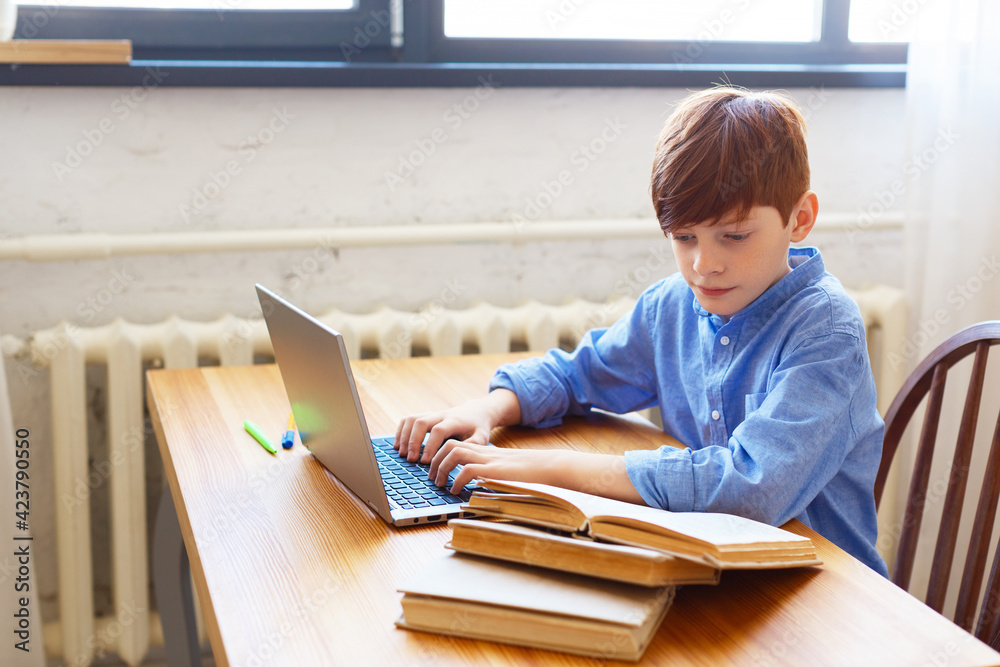 Teen boy doing homework online at home. He looks at the textbook and types a message with questions to the teacher. Teen boy doing homework online at home. He looks at the textbook and types a message with questions to the teacher.