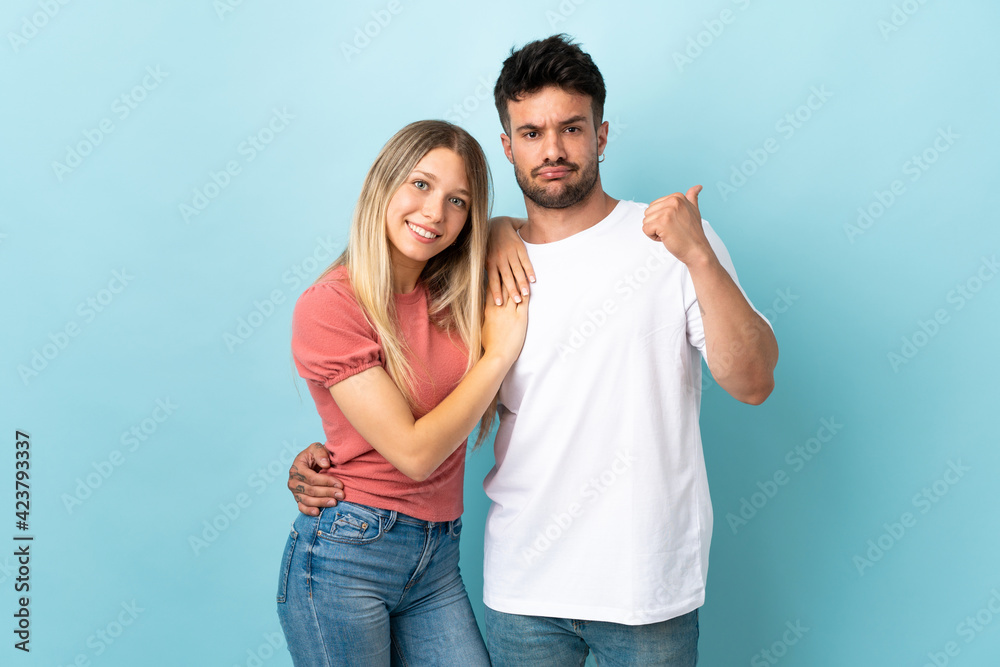 Young caucasian couple isolated on blue background frustrated and pointing finger to the side Young caucasian couple isolated on blue background frustrated and pointing finger to the side