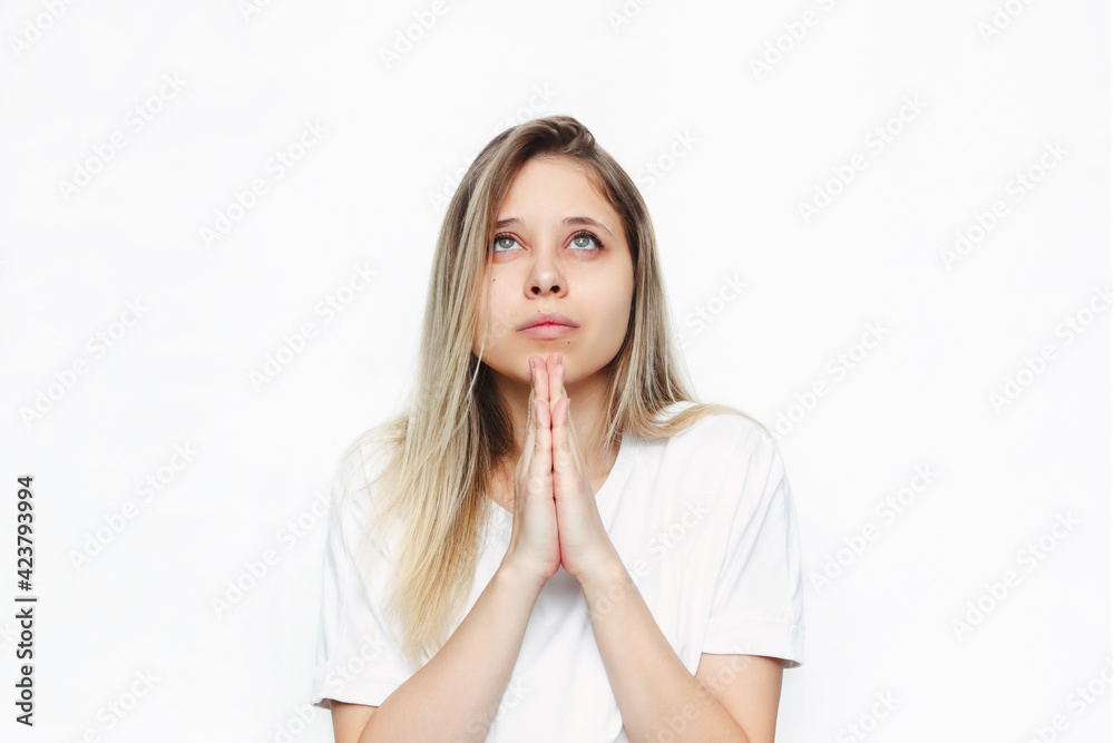 A young caucasian beautiful blonde woman in a white t-shirt prays with folded hands and looks up at the sky, thanks, making wish, asking help or forgiveness isolated on white background A young caucasian beautiful blonde woman in a white t-shirt prays with folded hands and looks up at the sky, thanks, making wish, asking help or forgiveness isolated on white background