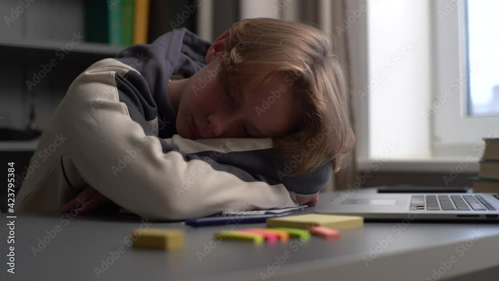 Close-up of tired pupil boy sleeping on workbook in front of laptop after online lesson via Internet near window. Concept of distance education at home during quarantine. Tracking shot in slow motion. Close-up of tired pupil boy sleeping on workbook in front of laptop after online lesson via Internet near window. Concept of distance education at home during quarantine. Tracking shot in slow motion.