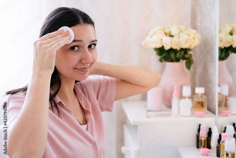 Cute caucasian woman touching face with cotton disk in bedroom. Beauty self care concept. Cute caucasian woman touching face with cotton disk in bedroom. Beauty self care concept.