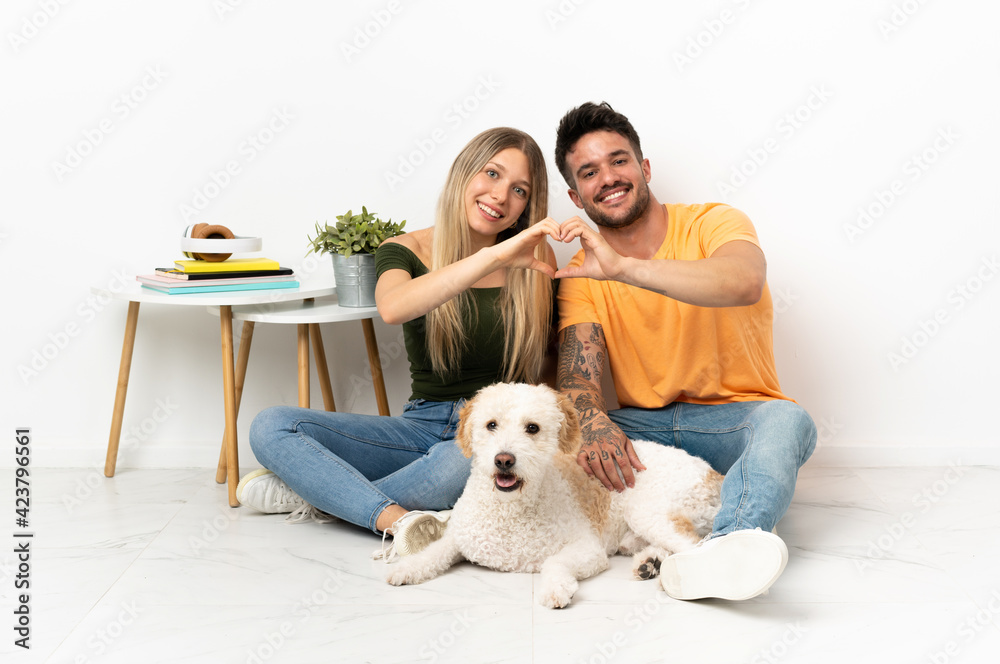 Young caucasian couple with dog staying at home making a heart with hands