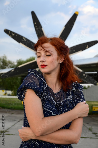 A beautiful red-haired girl in a blue dress with white polka dots stands no against the background of a small retro airplane