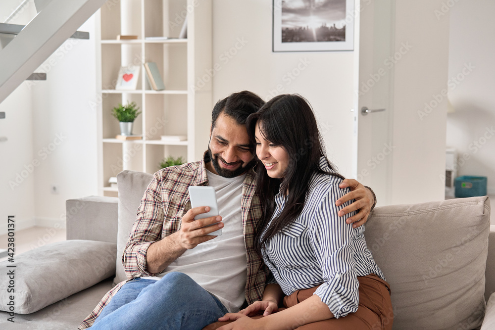Smiling young adult indian family couple embracing holding smartphone using mobile apps on cellphone watching social media videos, doing ecommerce shopping ordering online sitting on couch at home. Smiling young adult indian family couple embracing holding smartphone using mobile apps on cellphone watching social media videos, doing ecommerce shopping ordering online sitting on couch at home.