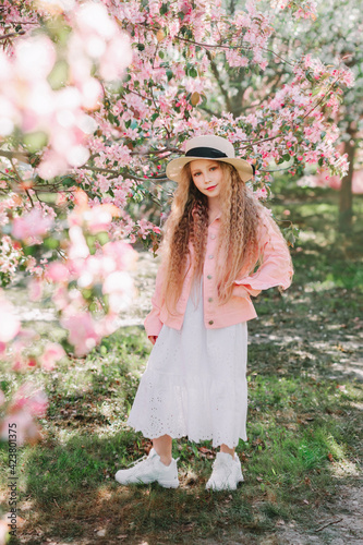 Beautiful longhaired blonde girl, She wearing white dress, pink jacket and straw and walking in apple blooming garden.