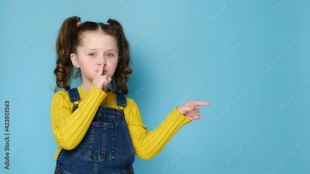 Emotional cute little girl holding finger on lips symbol of hush gesture of asking to be quiet, isolated over blue studio background with copy free space for advertisement. Silence or secret concept Emotional cute little girl holding finger on lips symbol of hush gesture of asking to be quiet, isolated over blue studio background with copy free space for advertisement. Silence or secret concept