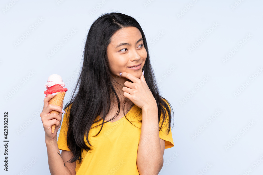 Teenager Asian girl with a cornet ice cream isolated on blue background thinking an idea and looking side