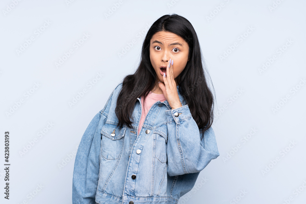 Teenager Chinese woman isolated on blue background whispering something with surprise gesture while looking to the side Teenager Chinese woman isolated on blue background whispering something with surprise gesture while looking to the side