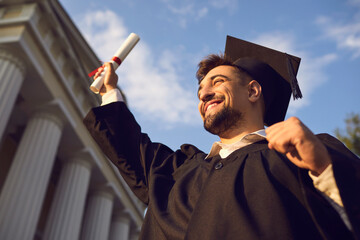 Low angle portrait of happy triumphant male graduate standing near university holding up diploma. From below of young handsome man proud of academic achievements celebrating college graduation